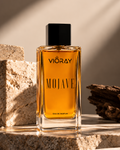 A bottle of Vioray Mojave Eau de Parfum with amber-colored liquid and a black cap, styled on a stone surface with textured blocks and driftwood under warm directional sunlight.
