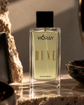 A rectangular glass bottle of Vioray Dune Eau de Parfum with a black cap, filled with pale golden liquid, standing on a stone surface with warm sunlight and shadow. Decorative rocks, bowls, and a textured background surround it.