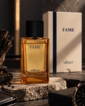 A bottle of Vioray FAME fragrance with amber liquid and a black textured cap stands on a stone slab beside its cream-colored box, styled with dark wood and a watch in a moody setting.
